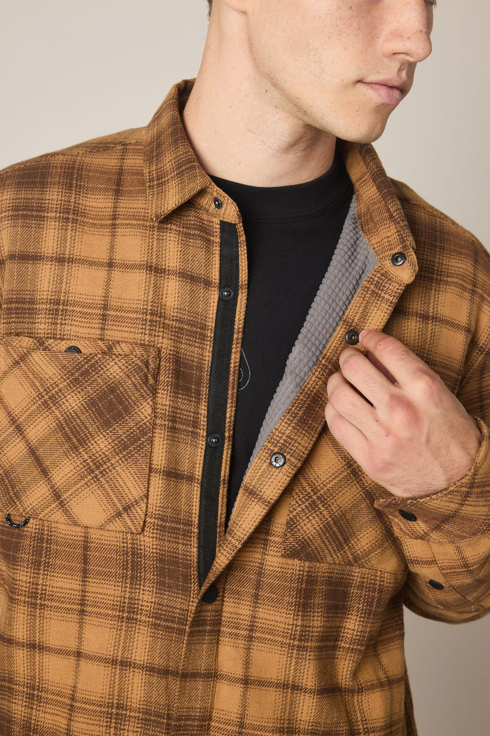 A man wearing the 686 Men's Heat Grid™ Flannel—a brown plaid shirt made from 100% polyester by 686—over a black T-shirt holds the flannel open with his right hand to reveal its soft, textured gray lining. His face is partially visible.