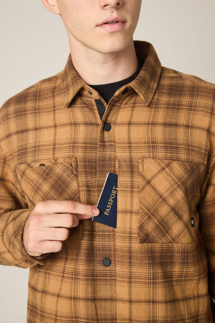A person wearing the 686 Men's Heat Grid™ Flannel by 686 places a blue passport into the chest pocket, with only the lower half of their face and upper torso visible.