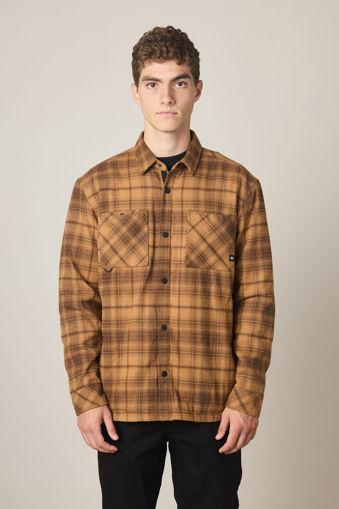 A person stands facing forward against a plain light background, wearing the 686 Men's Heat Grid™ Flannel by 686—a brown plaid shirt made from 100% polyester with two chest pockets—paired with black pants.