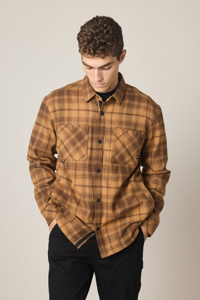A person with short curly hair wears the 686 Men's Heat Grid™ Flannel by 686 in a brown plaid pattern, featuring front pockets, while looking down with their hands in black pants pockets against a plain background.