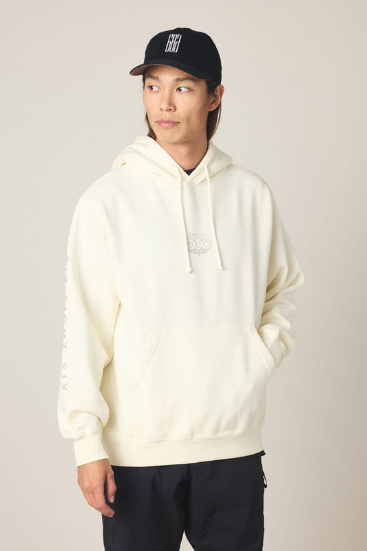 A person wearing the 686 Men's High Def Relaxed LS Hoody in cream, a cotton sweatshirt with a front pocket, and a black baseball cap looks to the side. This 686 men's sweatshirt features subtle text on the sleeve and a small design on the chest. The background is plain.
