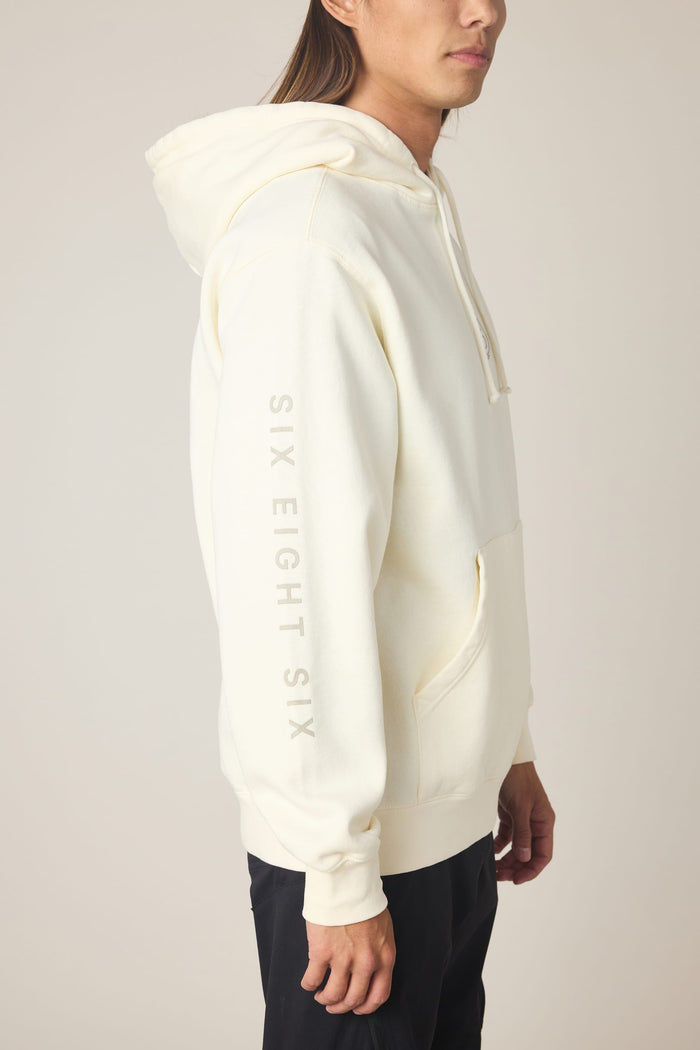 A person wearing the 686 Men's High Def Relaxed LS Hoody in off-white, featuring "SIX EIGHT SIX" printed vertically on the sleeve, stands in profile against a plain background.