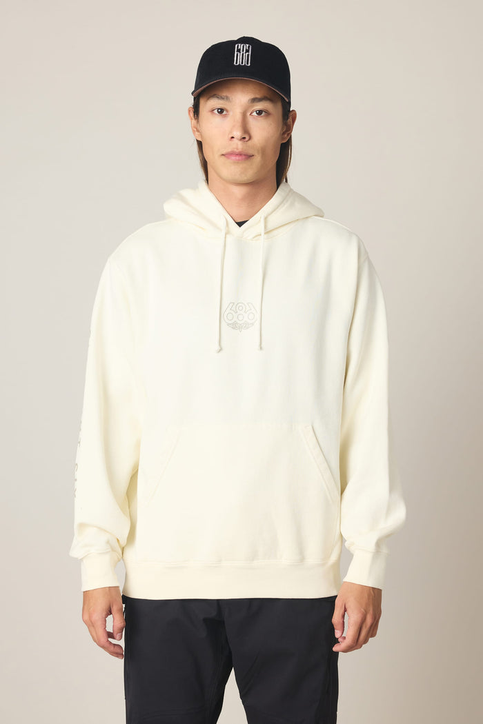 A person wearing the 686 Men's High Def Relaxed LS Hoody in cream, featuring a subtle chest logo, and a black baseball cap stands facing forward against a plain light background.