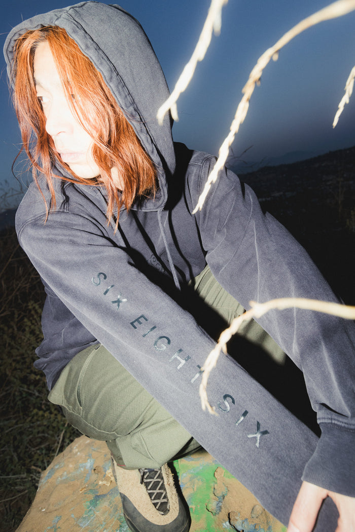 A person with red hair is sitting outdoors, wearing the 686 Men's High Def Relaxed LS Hoody and olive pants. The hoodie features the SIX EIGHT SIX logo on the sleeve. Bright light highlights their face, while dry branches partially block the view.