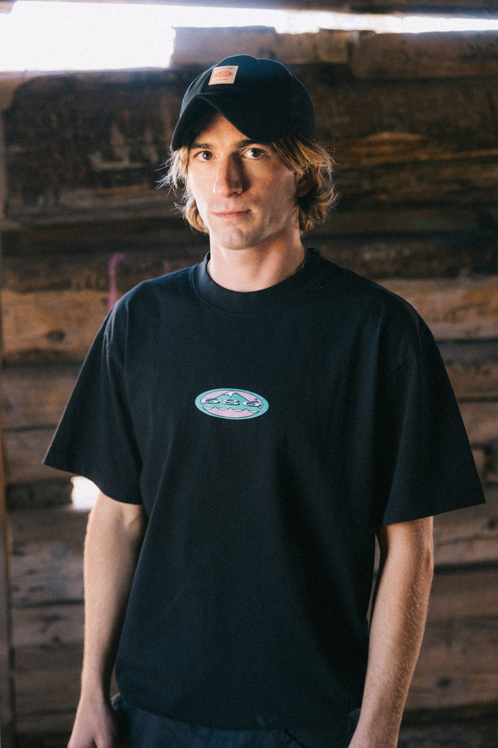 A young person with shoulder-length blond hair, wearing a black cap and the 686 Men's Jib Relaxed SS Tee by 686—an all-cotton classic black men’s t-shirt—stands indoors in front of a wooden wall, looking at the camera with a neutral expression.