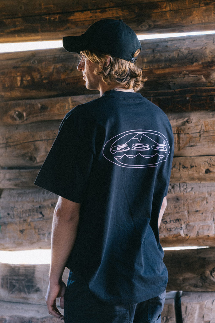 A person with wavy hair, wearing a black cap and the 686 Men's Jib Relaxed SS Tee by 686—which features a white graphic on the back—stands in front of a wooden wall, facing away from the camera.