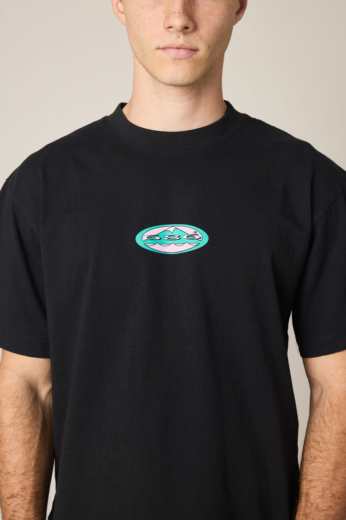 A person wearing the 686 Men's Jib Relaxed SS Tee by 686, a black 100% cotton t-shirt featuring a small, colorful oval graphic on the chest, stands against a plain background.