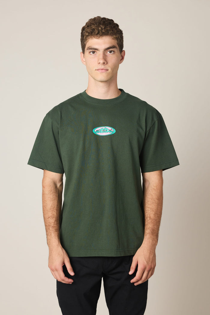 A young man with short curly hair wears the 686 Men's Jib Relaxed SS Tee, featuring a small, colorful oval logo on the chest, against a plain beige background. This stylish cotton t-shirt from 686 offers everyday comfort and effortless style.