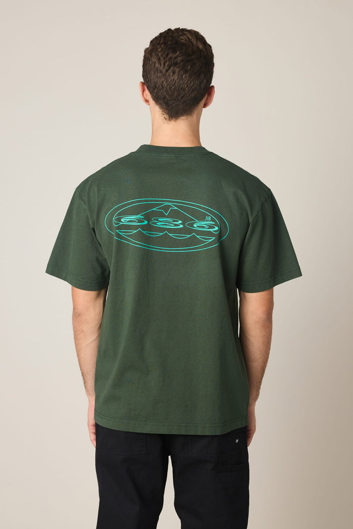 A person wearing the 686 Men's Jib Relaxed SS Tee in dark green, made from 100% cotton and featuring a blue oval graphic on the back, stands facing away from the camera against a plain background.