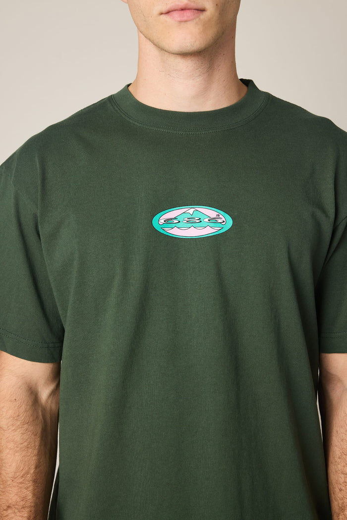 A person wearing the 686 Men's Jib Relaxed SS Tee in dark green, featuring a small, oval-shaped mountain and cloud graphic on the chest. The person's face is partially out of frame.