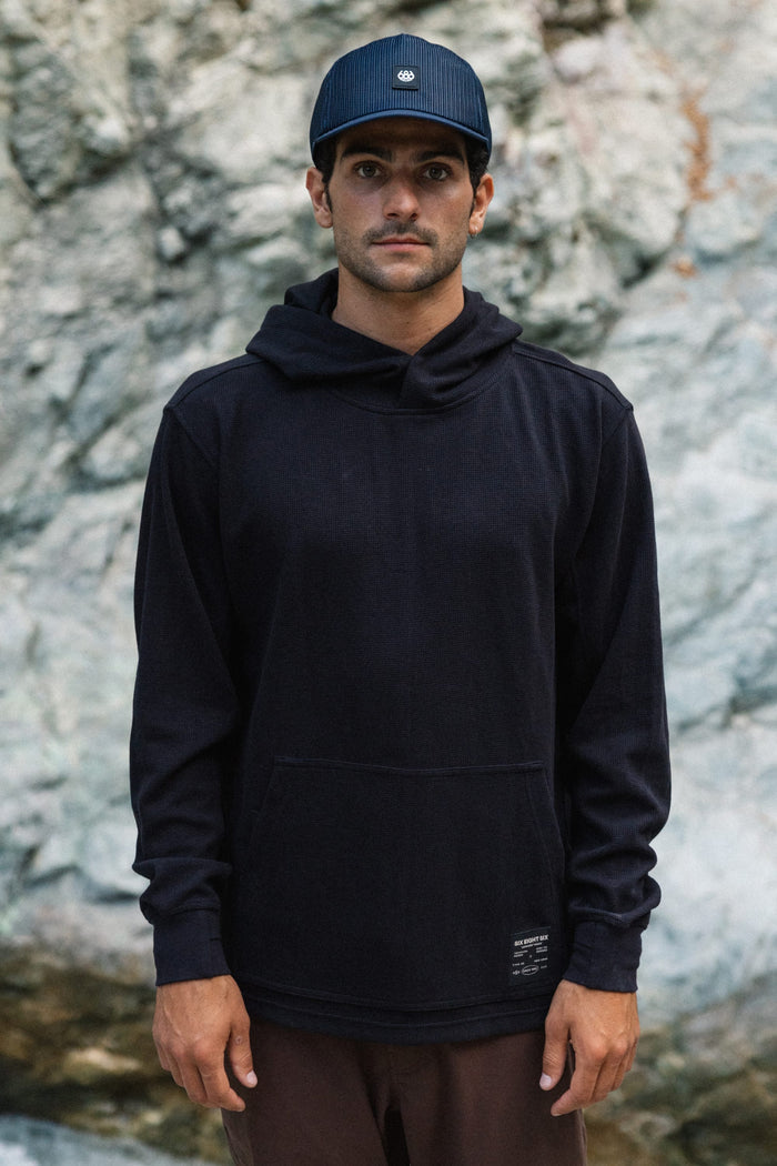 A man wearing the 686 Men's Let's Chill Waffle Hoody by 686 over a men's polyester t-shirt and a navy cap stands in front of a light-colored rocky background, looking directly at the camera with a neutral expression.