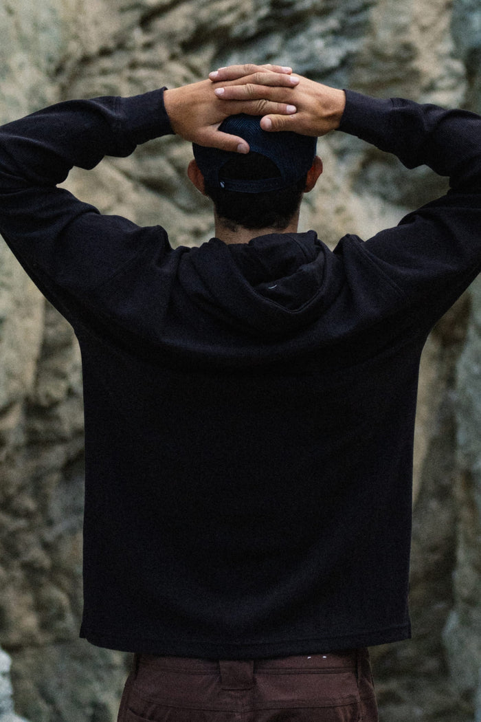 Wearing the 686 Men's Let's Chill Waffle Hoody and a cap over a men's polyester t-shirt, an individual stands with their back to the camera, hands clasped on their head, against a backdrop of rugged rocks.