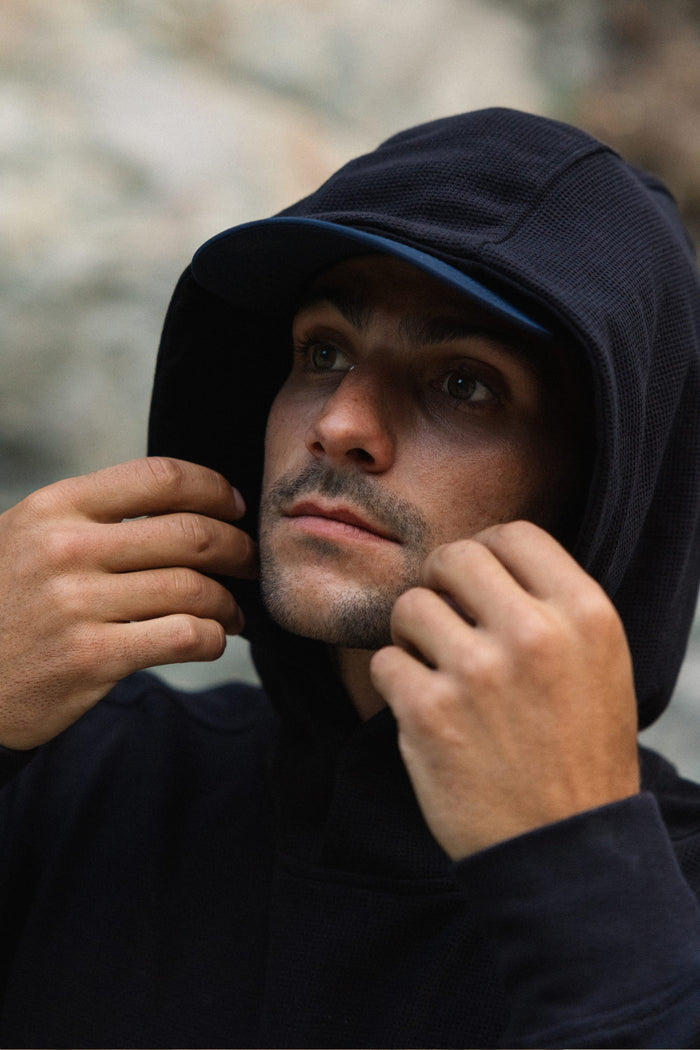 A person wearing the 686 Men's Let's Chill Waffle Hoody by 686 over a men's polyester t-shirt and cap looks up while adjusting the hood with both hands, with a blurred background that draws focus to their face and hands.