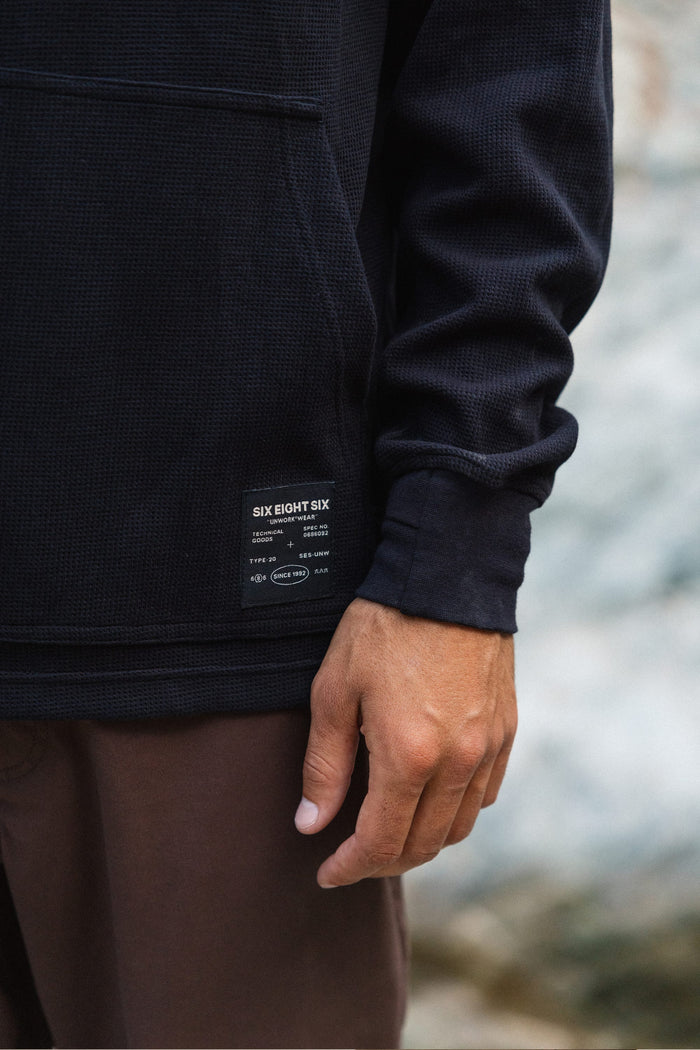 A person wearing the 686 Men's Let's Chill Waffle Hoody in black, featuring a visible 686 label, pairs it with brown pants while standing outdoors with one hand relaxed at their side—effortlessly layering the look as you might with a classic men's polyester t-shirt.