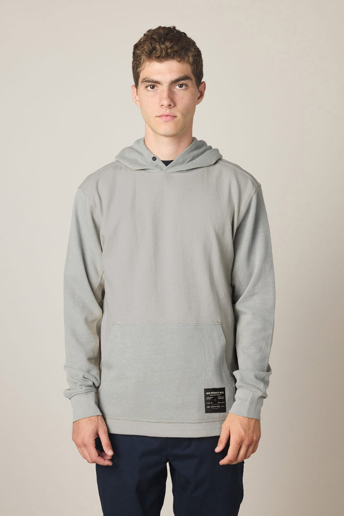 A young man stands facing forward, wearing the 686 Men's Let's Chill Waffle Hoody in light gray with a front pocket and dark pants over a classic men's polyester t-shirt, set against a plain light background.