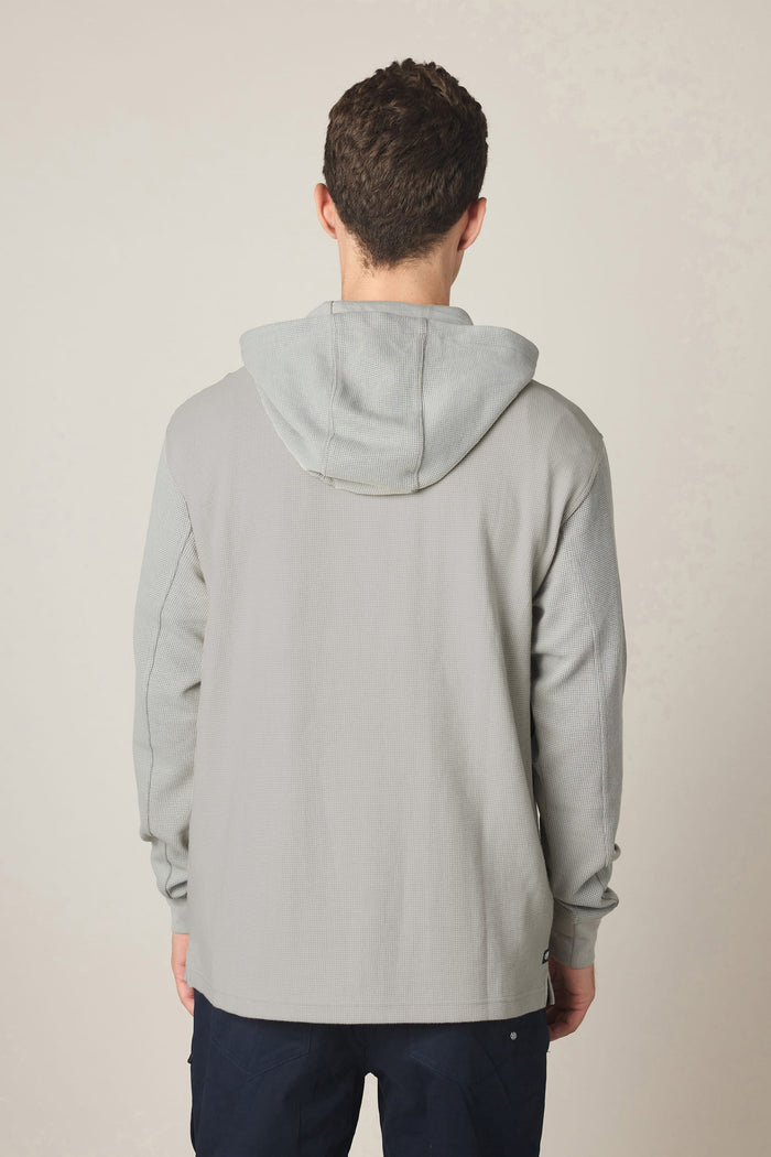A person with short brown hair is standing facing away from the camera, wearing the 686 Men's Let's Chill Waffle Hoody layered over a men's polyester t-shirt and dark pants against a neutral background.
