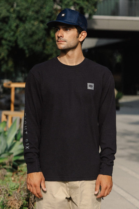 A man wearing the 686 Men's Let's Chill Waffle LS Crew by 686 and a blue cap stands outdoors, looking to the side. Green plants and a building are visible in the background.