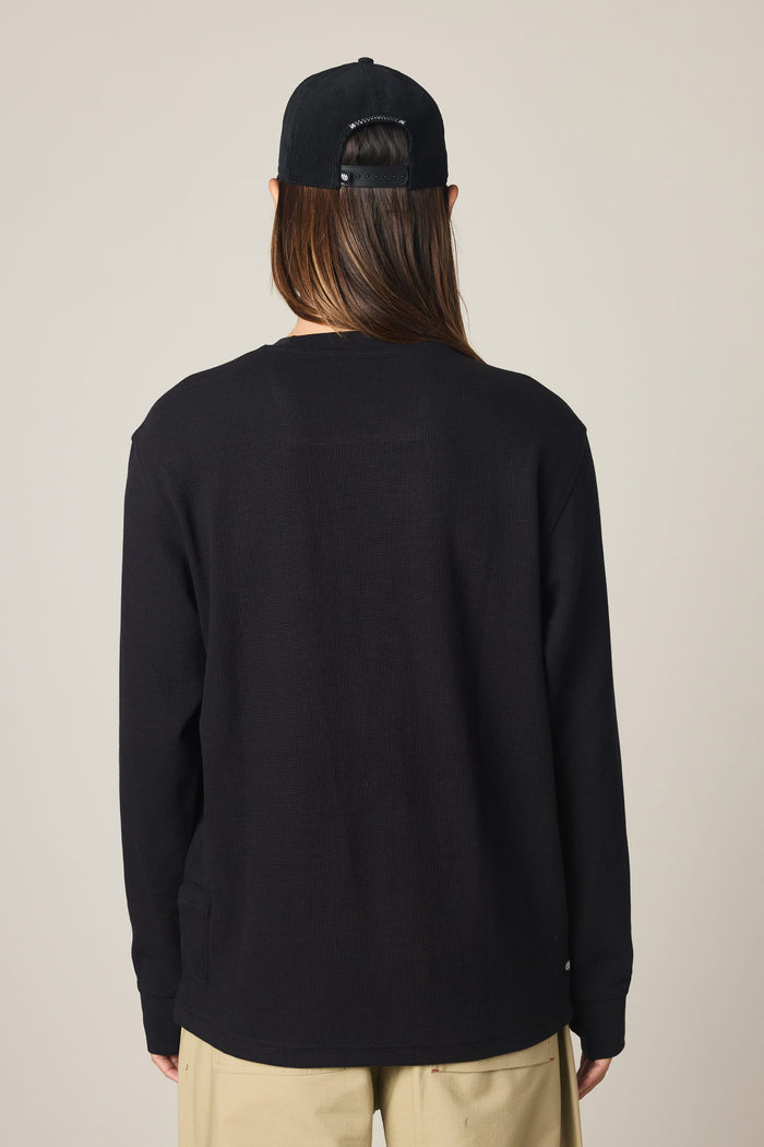 A man is shown from the back against a plain light background, wearing the 686 Men's Let's Chill Waffle LS Crew in black, paired with beige pants and a black cap.