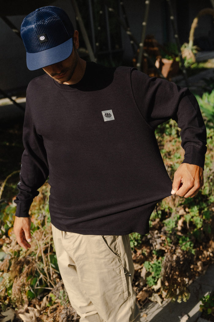 A person stands outdoors amid greenery and sunlight, wearing a navy blue cap, beige pants, and the 686 Men's Let's Chill Waffle LS Crew by 686 in black polyester. They are holding out the side of their shirt.