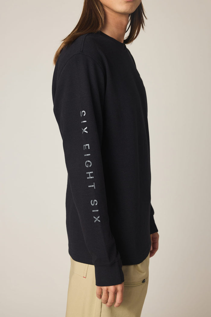 A person wearing the 686 Men's Let's Chill Waffle LS Crew in black with "686" written down the sleeve, paired with tan pants, stands sideways against a plain background.