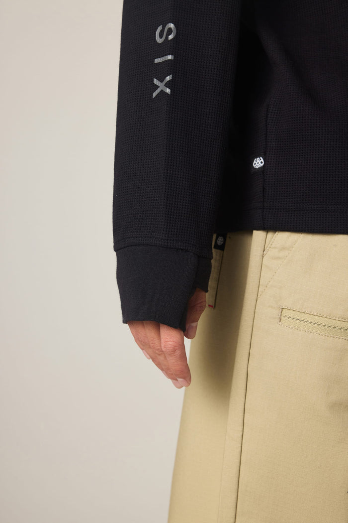 A person is pictured from the waist down, wearing the 686 Men's Let's Chill Waffle LS Crew— a black long-sleeve polyester t-shirt by 686 with lettering on the sleeve—paired with beige pants, showing their hand and part of their pants pocket.