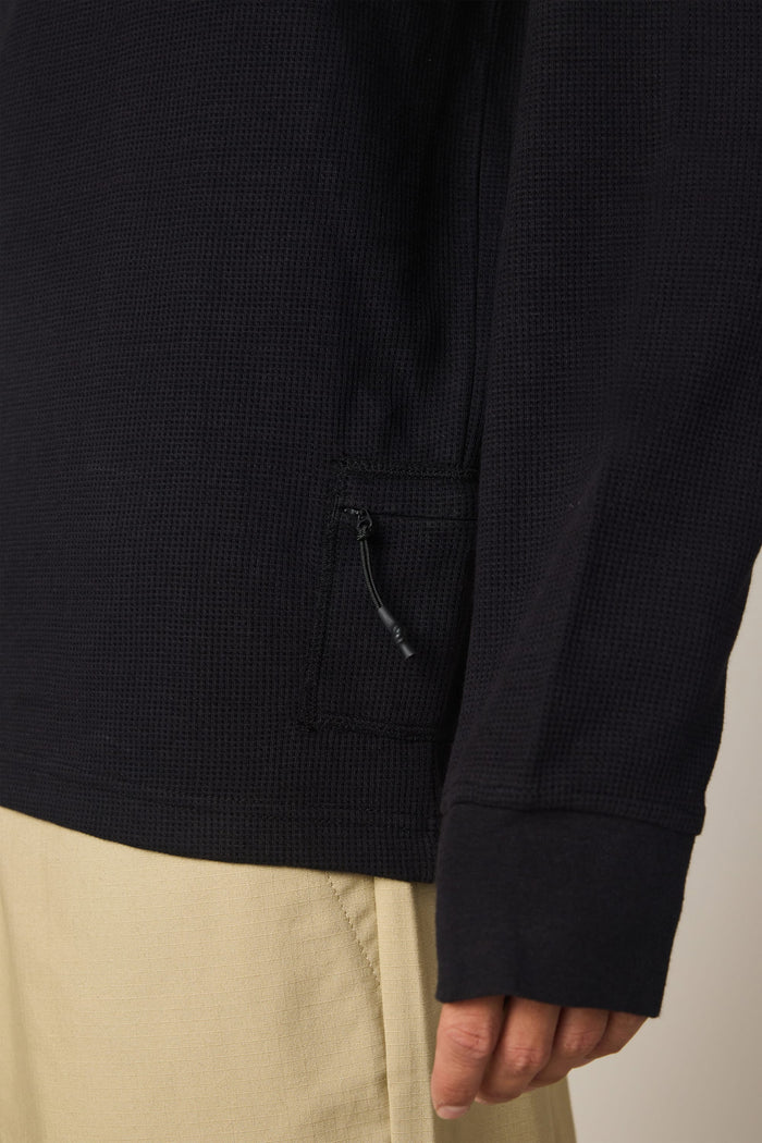 A close-up of a person wearing the 686 Men's Let's Chill Waffle LS Crew in black—a textured long-sleeve polyester t-shirt with a small zippered pocket—paired with light beige pants. Only the lower half of the shirt and a hand are visible.