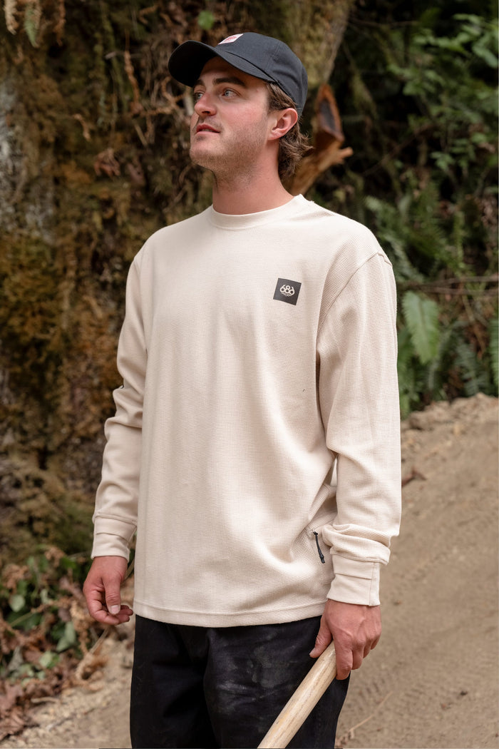 A man wearing the 686 Men's Let's Chill Waffle LS Crew in beige and a black cap stands outdoors holding a shovel, looking up. Trees, plants, and dirt can be seen in the background.
