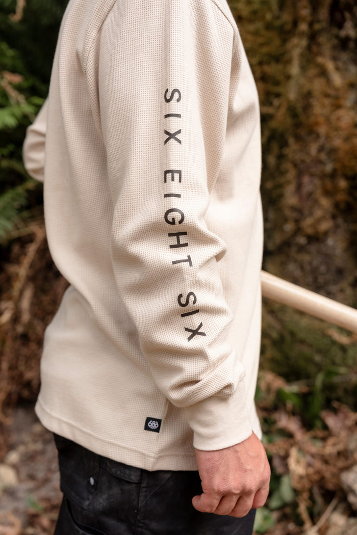 A person wearing the 686 Men's Let's Chill Waffle LS Crew—a beige polyester t-shirt by 686 with “SIX EIGHT SIX” printed vertically on the sleeve—stands outdoors, facing sideways against a backdrop of blurred greenery and natural elements.