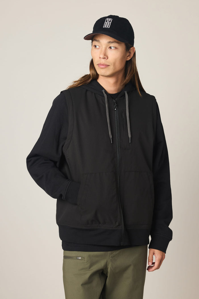 A person wearing a black cap, olive-green pants, and the 686 Men's Merge Quilted Hoody by 686—a black polyester jacket with gray drawstrings—stands against a plain light background, looking slightly to the side with hands in the jacket pockets.