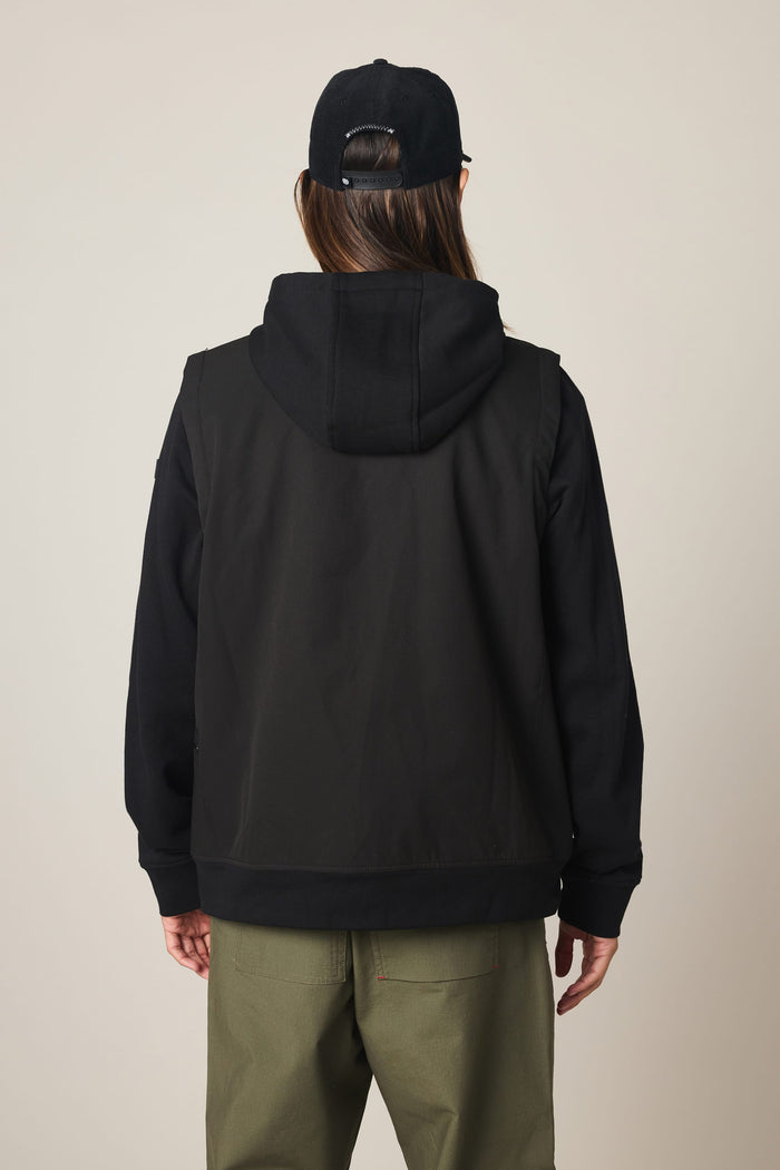 A person stands facing away from the camera, wearing the 686 Men's Merge Quilted Hoody layered under a men’s polyester jacket, paired with green pants and a black baseball cap.