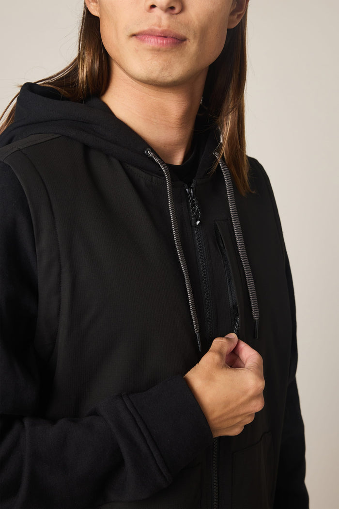 A person with long brown hair, shown from the shoulders up against a plain, neutral background, is wearing the 686 Men's Merge Quilted Hoody by 686 and holding the zipper near their chest.