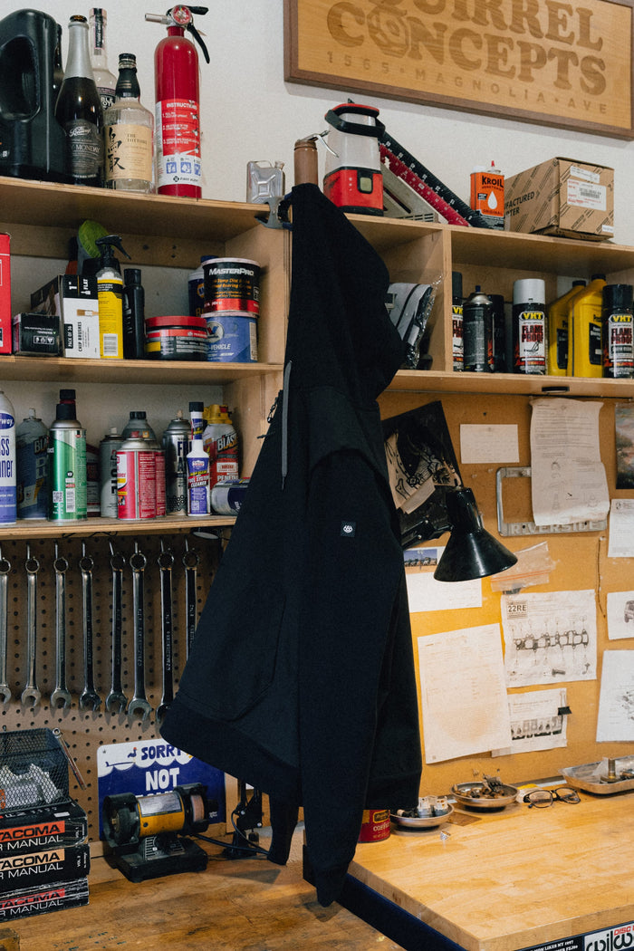 A 686 Men's Merge Quilted Hoody hangs on a hook in a cluttered workshop, surrounded by shelves of tools, oil cans, a fire extinguisher, and papers pinned above a wooden workbench.