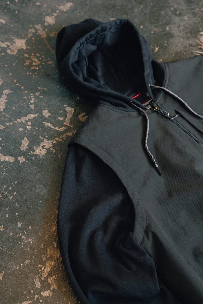 A 686 Men's Merge Quilted Hoody in black, featuring polyester knit construction and a zipper, is laid flat on a textured, worn concrete surface. Only the upper half of the hoody and its hood are visible.