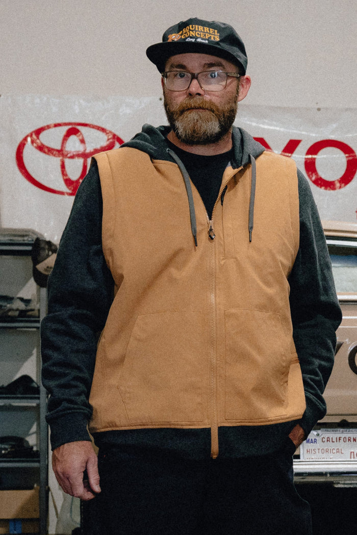 A man with a beard and glasses wears the 686 Men's Merge Quilted Hoody under a tan men's polyester jacket and a black cap. He stands indoors in front of a Toyota banner, with shelves and automotive items visible behind him.