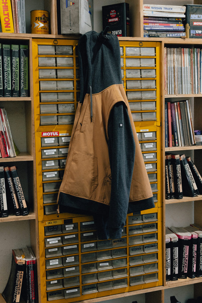 A 686 Men's Merge Quilted Hoody in tan and black hangs on a yellow cabinet with many small drawers, surrounded by bookshelves filled with books and magazines.