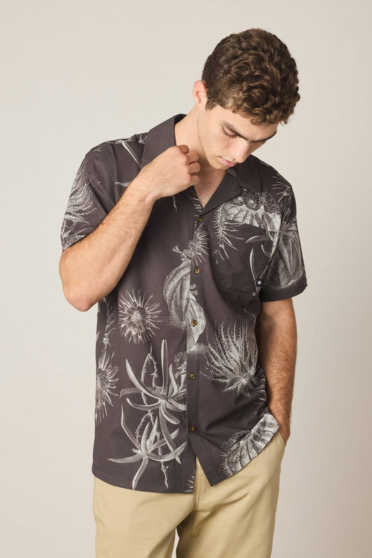 A man with short curly hair wears the 686 Men's Nomad Perforated Button Down Shirt, featuring a white botanical print, and light khaki pants, looking down against a plain background with one hand on his head and the other in his pocket.