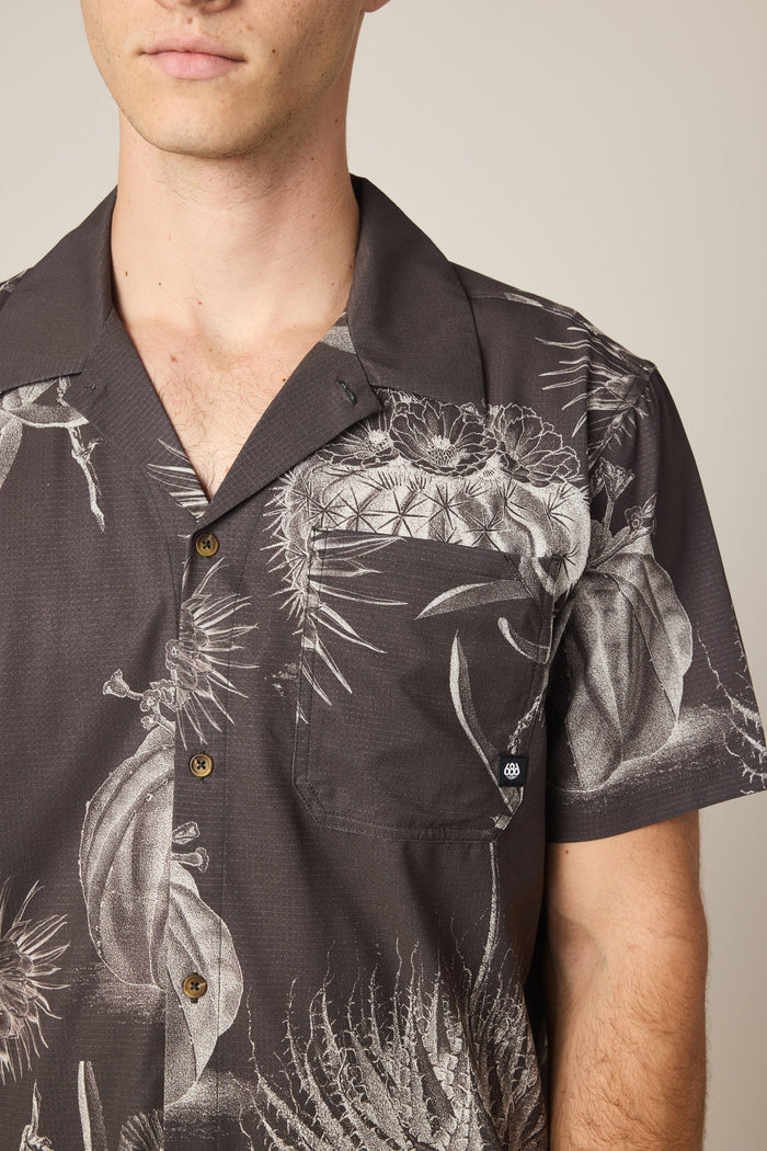 A man wears the 686 Men's Nomad Perforated Button Down Shirt‚Äîa dark, lightweight shirt with a white botanical and cactus print, chest pocket, and brown buttons. Only his upper torso and part of his face are visible.