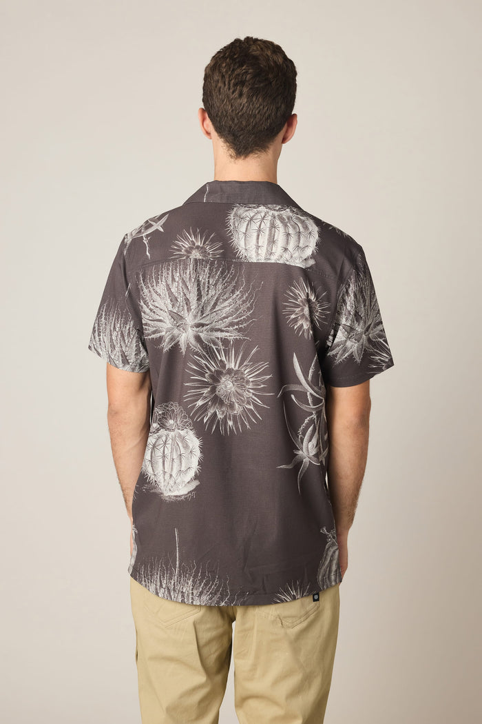 A man, seen from behind against a plain background, wears the 686 Men's Nomad Perforated Button Down Shirt by 686‚Äîa dark lightweight shirt featuring a white cactus print‚Äîpaired with light-colored pants.