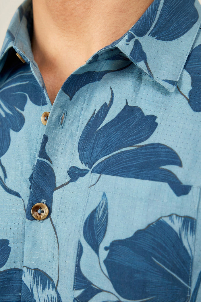 Close-up of a person wearing the 686 Men's Nomad Perforated Button Down Shirt in light blue with a dark blue floral pattern and brown buttons. This lightweight, airy shirt by 686 features a collar and is partially unbuttoned at the top.