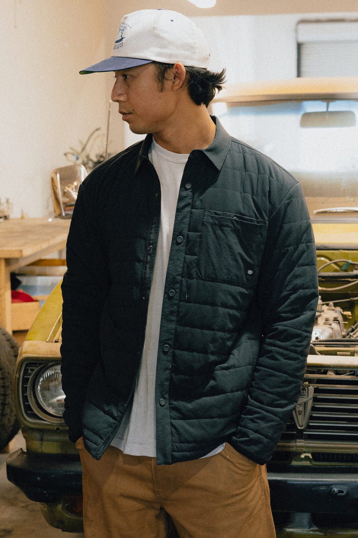 A man in a white cap, wearing the 686 Men's Thermaldry™ Insulated Shacket by 686 over a white shirt and brown pants, stands in front of an old yellow car in a garage, looking to his left.