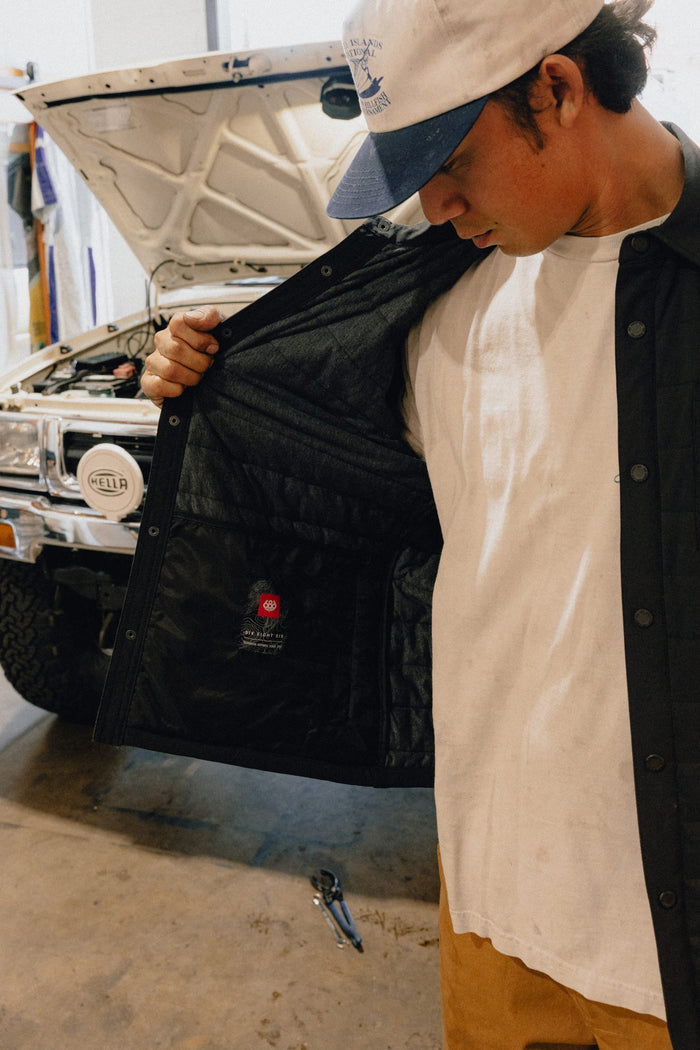 A person stands next to a white vehicle with its hood open, holding open their 686 Men's Thermaldry™ Insulated Shacket to reveal the quilted interior lining and the 686 label inside, while looking down at it.