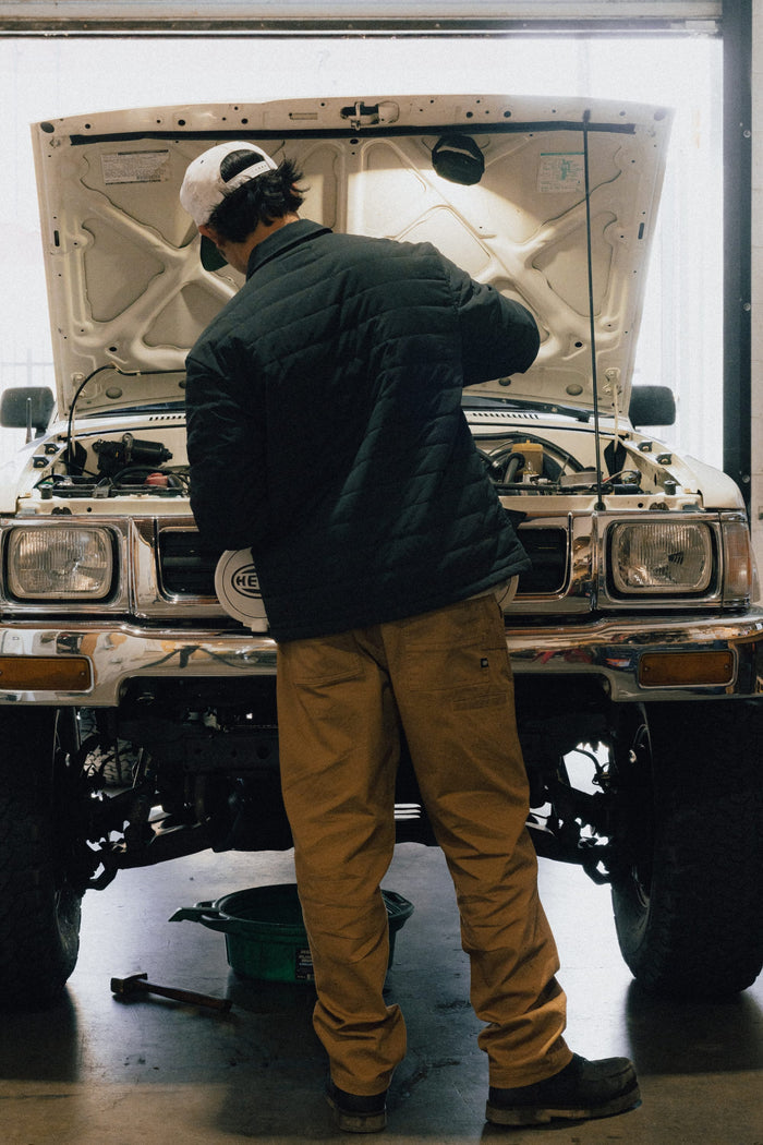 Wearing a 686 Men's Thermaldry™ Insulated Shacket, a person in tan pants works under the hood of a white SUV in a garage, with tools and a green container on the floor.