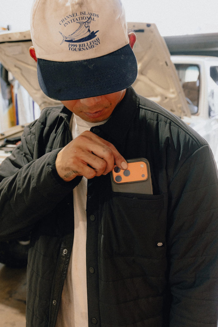 A person wearing a white cap and the 686 Men's Thermaldry™ Insulated Shacket places an orange iPhone in their chest pocket, with a car and an open hood in the background.