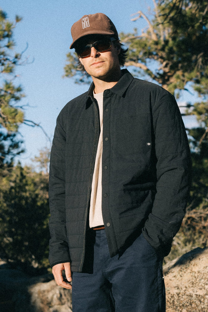 A man wearing sunglasses, a brown cap, the 686 Men's Thermaldry™ Insulated Shacket by 686, and dark pants stands outdoors near trees and rocks, looking at the camera.
