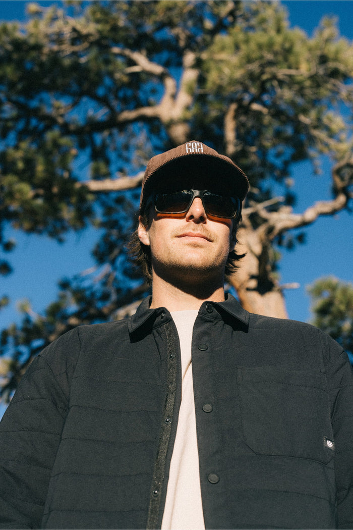 A person wearing a brown hat, sunglasses, and the 686 Men's Thermaldry™ Insulated Shacket by 686 stands outdoors in front of a tree with a bright blue sky in the background.