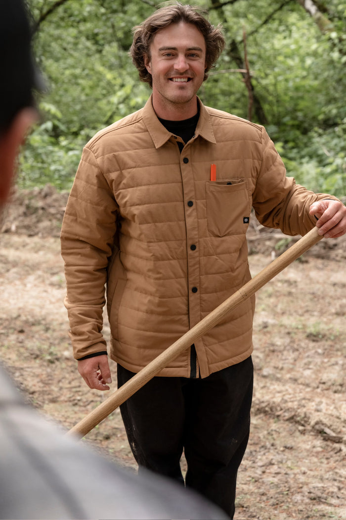 A person in a tan 686 Men's Thermaldry™ Insulated Shacket smiles while holding a wooden tool outdoors, with trees in the background and another person partly visible in the foreground.