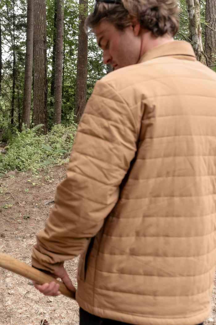 Wearing the 686 Men's Thermaldry™ Insulated Shacket, a person stands in a forested area holding a wooden stick, facing slightly away from the camera.