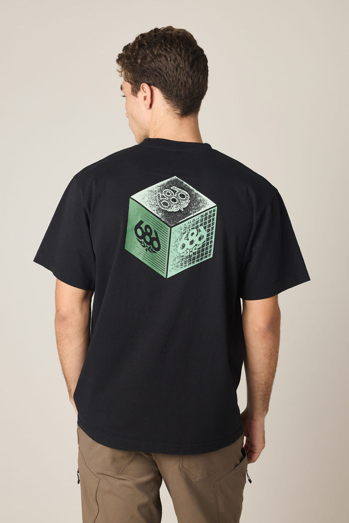 A person with short brown hair, seen from behind, wears the 686 Men's Unit Relaxed SS Tee—a black 100% cotton T-shirt by 686 featuring a green and white cube graphic with skull designs on the back. He is also wearing brown pants.