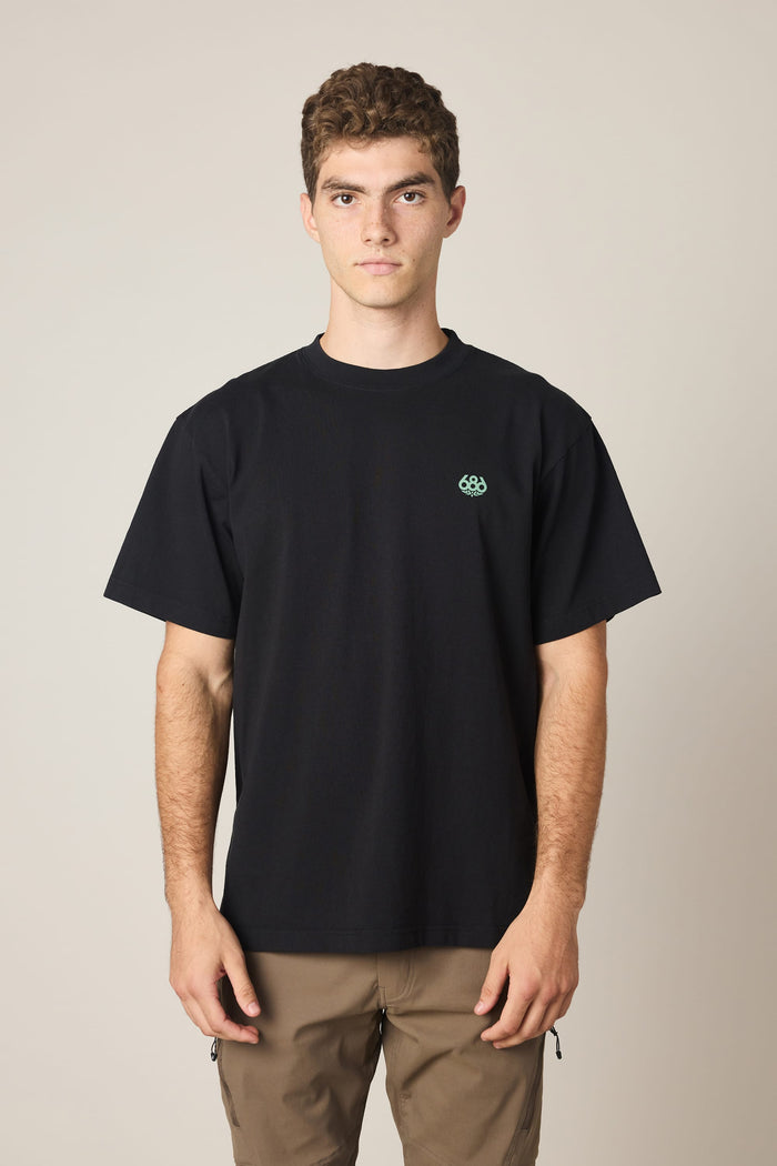 A young man with short curly hair wears the 686 Men's Unit Relaxed SS Tee, featuring a small green logo on the left chest, paired with light brown pants. He stands against a plain light background, looking directly at the camera.