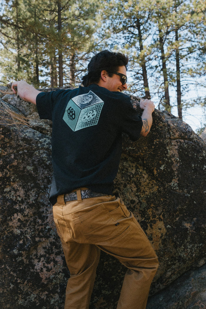 Wearing sunglasses, brown pants, and the 686 Men's Unit Relaxed SS Tee from 686—a black 100% cotton t-shirt featuring a green geometric design—a person climbs a large rock outdoors surrounded by tall pine trees.